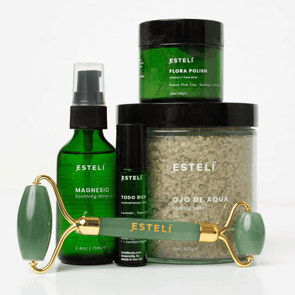 A selection of Esteli Natural skincare products including Magensio Mineral Sleep Spray, Todo Bien Aromatherapy wand, Ojo de Agua Bath Salts, Flora Polish Vitamin C Mask, and Aventurine Face Roller