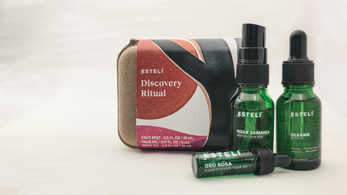Three green glass bottles—Rosa Jamaica Face Mist, Verano Body Oil, and Oro Rosa Face Oil—arranged beside ESTELÍ Discovery Ritual packaging.