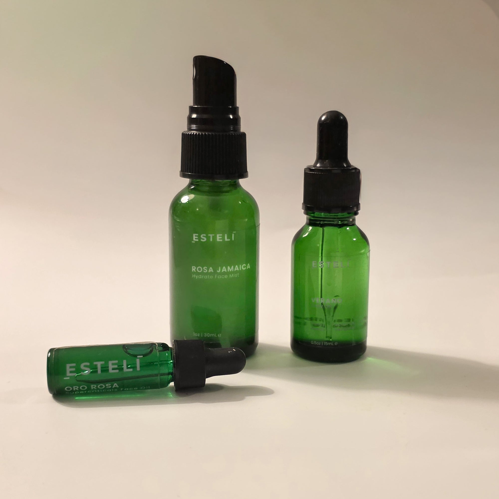 ESTELÍ Discovery Ritual featuring Rosa Jamaica Hydrating Face Mist, Oro Rosa Supercriticals Face Oil, and Verano Body Oil arranged on a warm cream background.