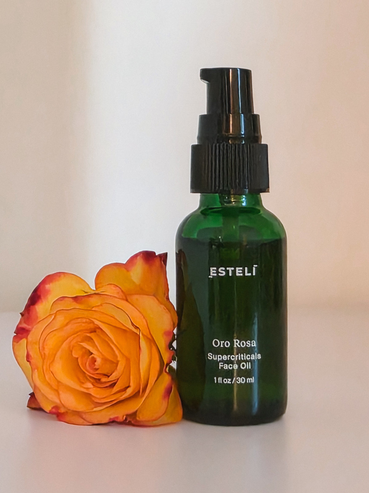 ESTELÍ Oro Rosa Supercriticals Face Oil in a green glass bottle beside a golden-orange rose with deep red petal edges.
