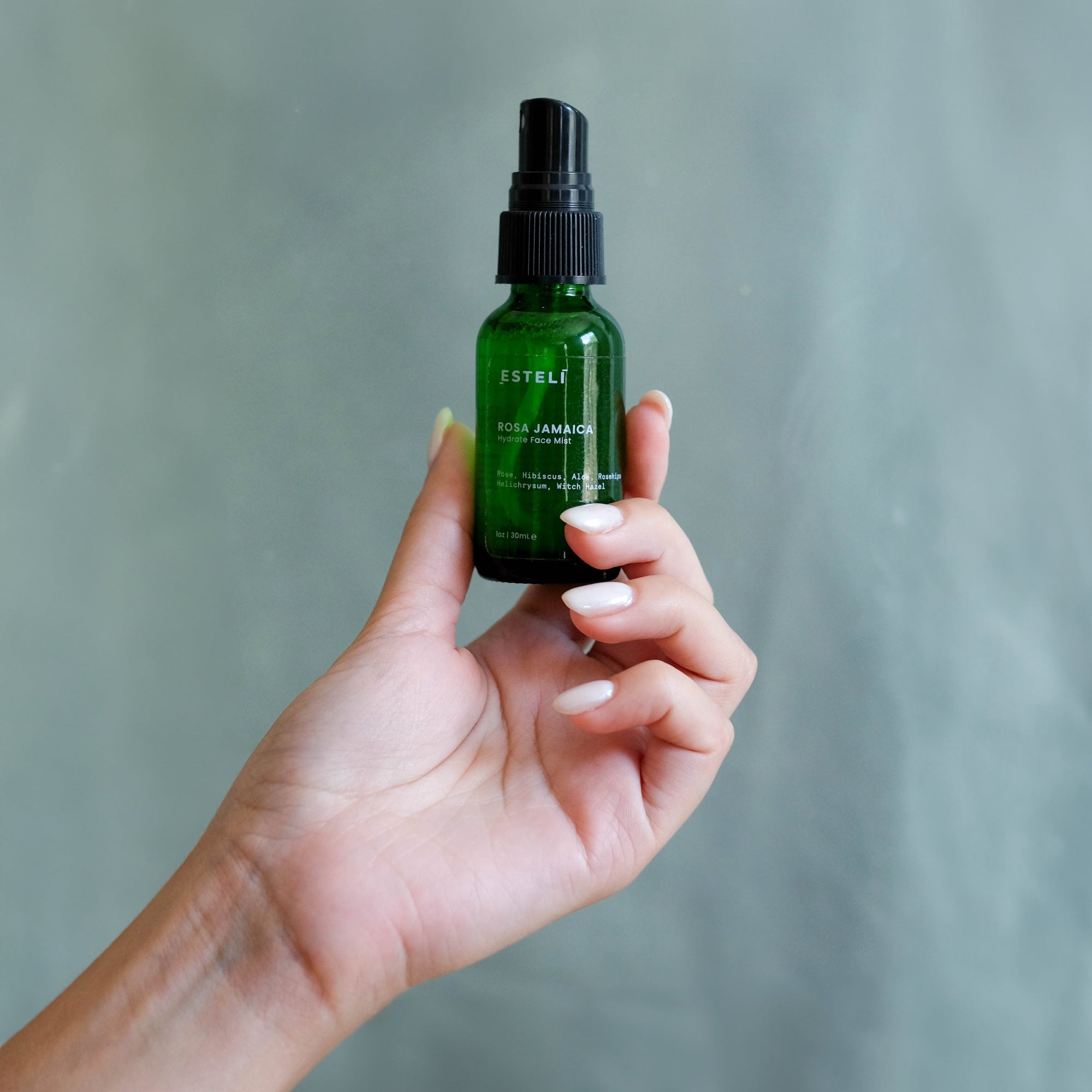 Hand holding ESTELÍ Rosa Jamaica Hydrating Face Mist mini in green glass