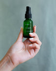 Hand holding ESTELÍ Rosa Jamaica Hydrating Face Mist mini in green glass
