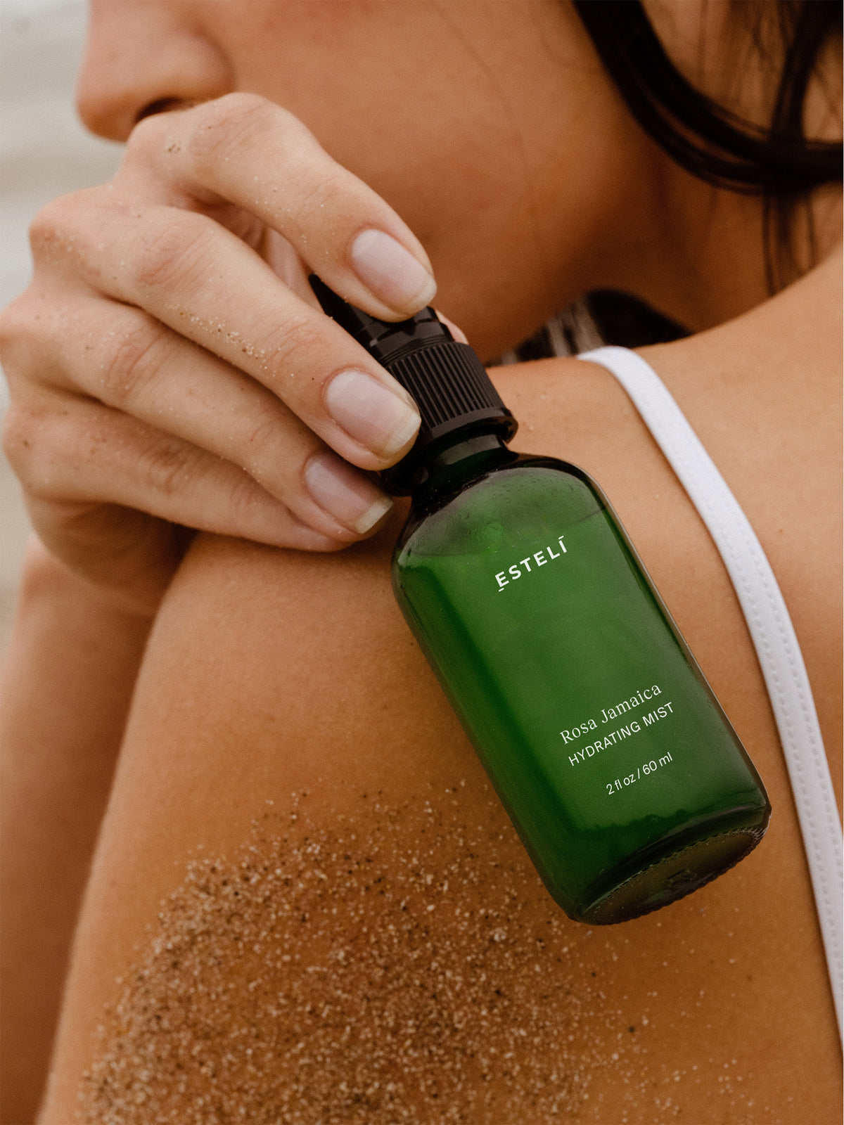Woman holding ESTELÍ Rosa Jamaica Hydrating Mist near her face, with sand on her skin.