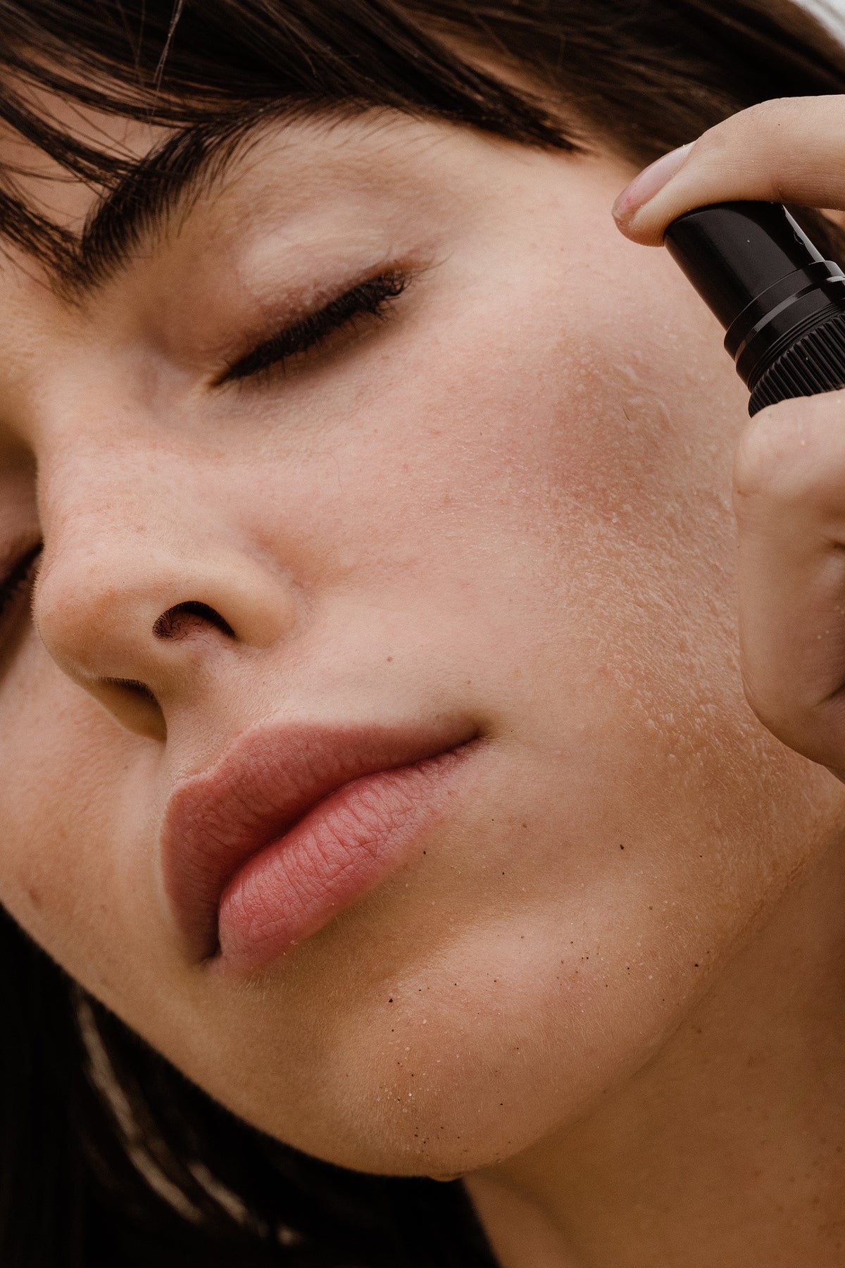 Woman with eyes closed applying ESTELÍ Rosa Jamaica Hydrating Face Mist, with fine droplets visible on her cheek.