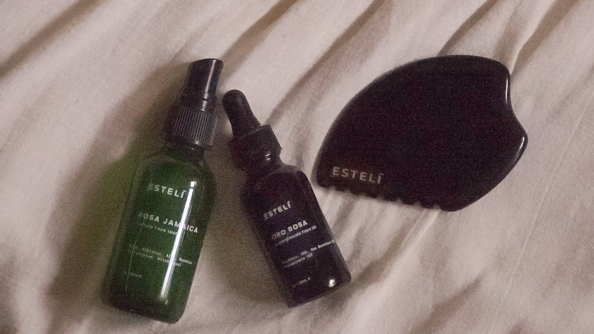 Esteli Rosa Jamaica botanical facial mist, Oro Rosa face oil, and black obsidian gua sha skincare ritual set on neutral linen